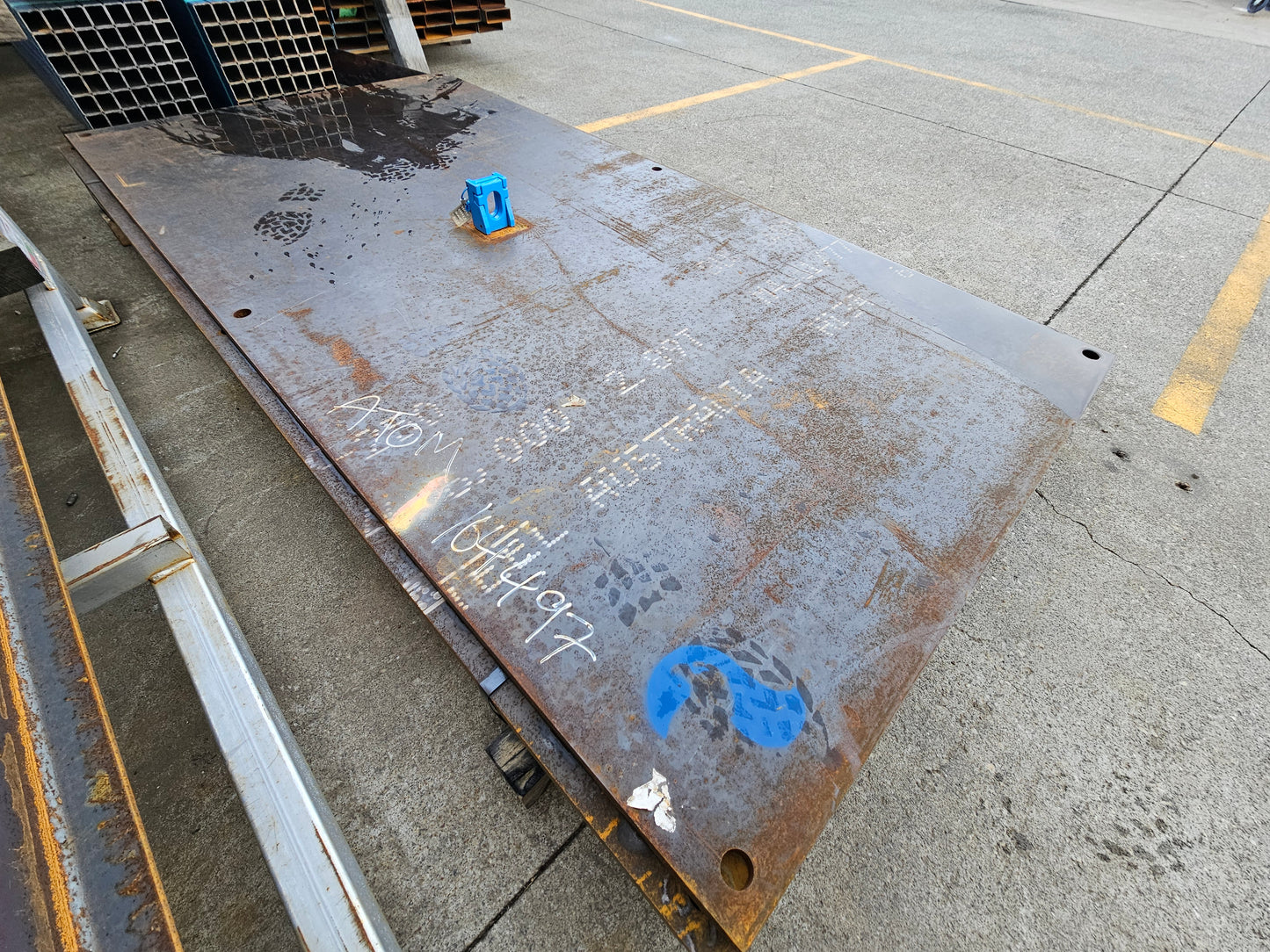 Yellow heavy duty steel road plate with a lifter and multiple fixing holes, placed on a concrete surface.