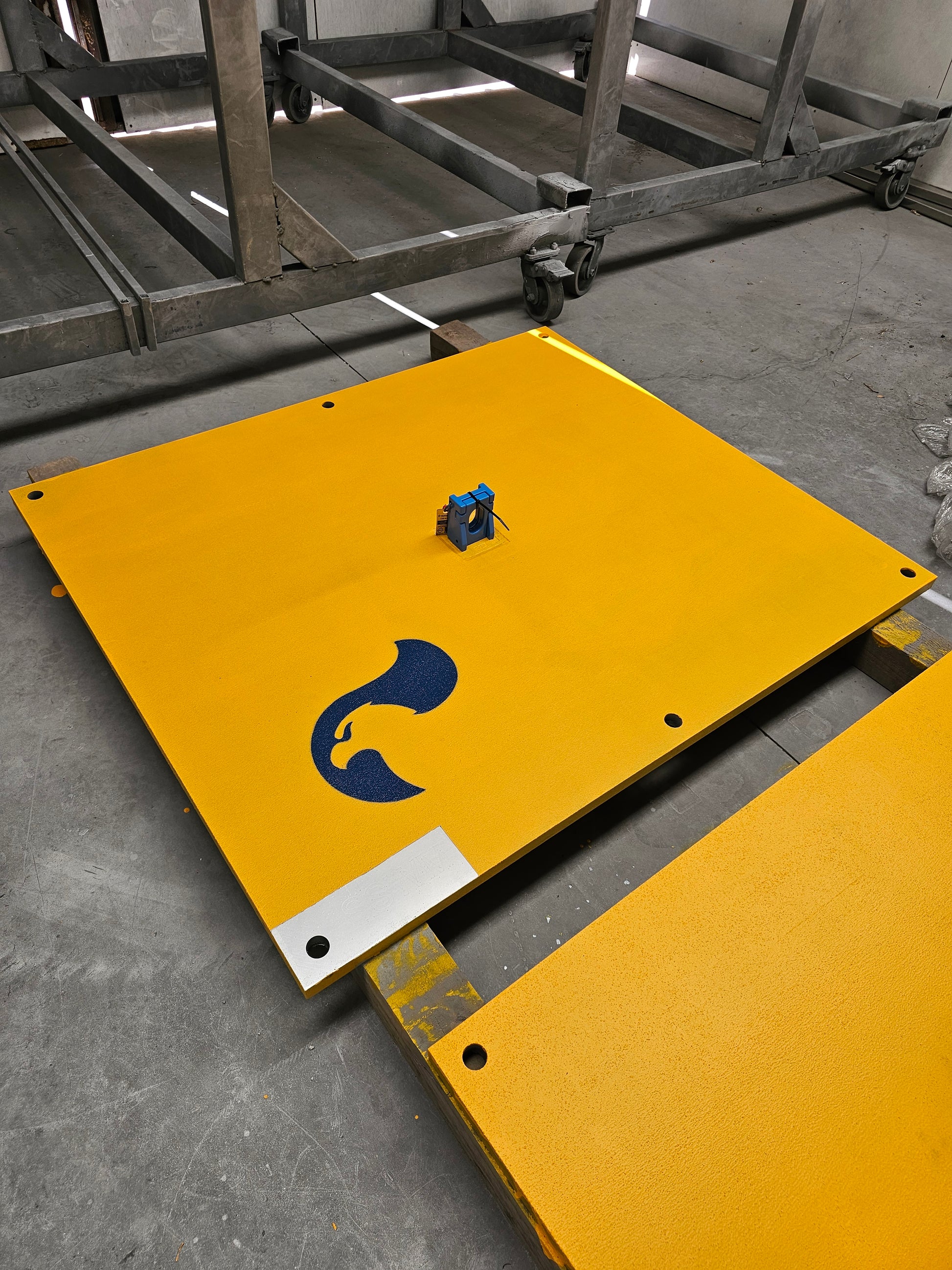 Yellow anti-slip coated RAPTA steel road plates laid out on a workshop floor. Shown with optional lifter. Custom sizes available. 
